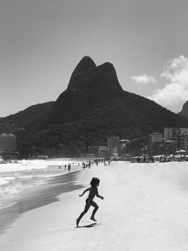 A black and white photo of a child running from the ocean on the sand with the dark silhouette of a hill in the background