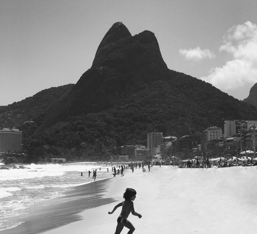 A black and white photo of a child running from the ocean on the sand with the dark silhouette of a hill in the background